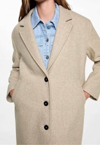 Béžový vlněný kabát s přizpůsobeným střihem, špičatými klopami a velkými černými knoflíky. Má boční kapsy a je vrstvený přes světle modrou džínovou košili.
