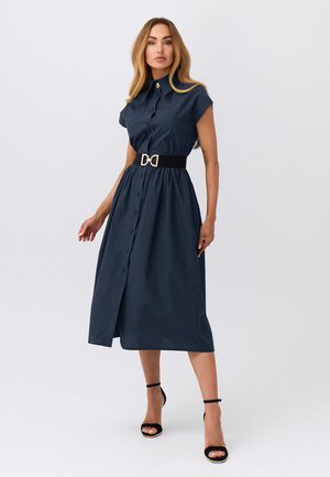 Kobieta w granatowej sukience midi zapinanej na guziki z krótkimi rękawami, czarnym paskiem ze złotą klamrą i czarnych szpilkach z paskiem wokół kostki, stojąca na białym tle.