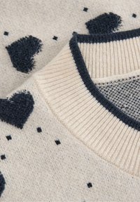 Strikket sweater i creme med marineblå accenter. Har teksturerede hjerte mønstre og små prikker. Ribkantet krave tilføjer detaljer og kontrast.