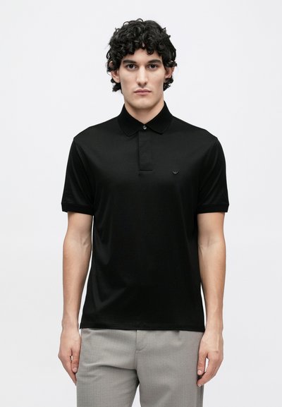 Melna polo krekls ar īsām piedurknēm, apkakli un smalku logo uz krūtīm. Gluda auduma ar pieguļošu dizainu, kopā ar gaiši pelēkām biksēm.