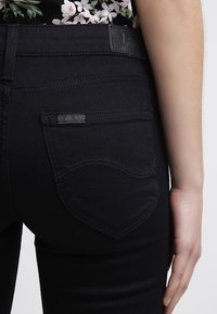 Jeans de mezclilla negra con un bolsillo trasero visible con costuras curvas y un parche de cuero en la cinturilla. Textura suave.