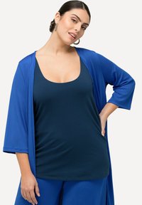 Modrý kimono-styl cardigan přes tmavě modrý tílko s kulatým výstřihem. Hladká tkanina s volným střihem a rukávy do lokte.