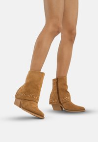 Botas de tobillo de gamuza color arena con un dobladillo doblado, adornadas con tachuelas decorativas y un tacón bajo, que cuentan con un cierre de cremallera en la parte trasera.
