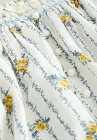 Vêtement en lin blanc avec des rayures florales bleues et une rose jaune, comprenant des boutons recouverts de tissu et une taille élastique froncée.