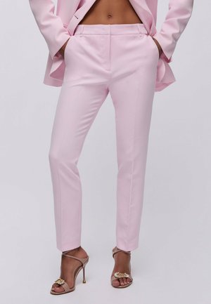 Mujer vistiendo pantalones sastre en rosa pastel y blazer a juego, combinados con sandalias beige de tacón alto que tienen hebillas decorativas.