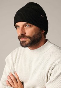 Zwarte gebreide beanie met ribbelstructuur en logotag, gedragen bij een lichtgekleurde ribbeltrui. Close-up van de gelaatskenmerken.