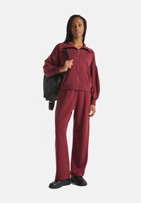 Bordeaux ritsjack met oversized mouwen en bijpassende relaxte broek, gecombineerd met een zwarte rugzak en zwarte schoenen.