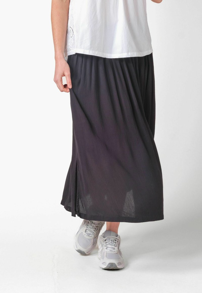 Femme portant une jupe midi plissée noire, un haut blanc et des baskets blanches, debout avec un pied légèrement avancé sur un fond blanc.