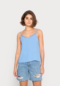 Camisola sem mangas de tom azul claro com rendas ao longo do decote em V, combinada com calções de ganga desgastados com bordas desfiadas e rasgos.