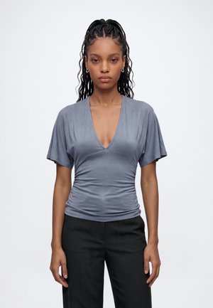 Femme aux cheveux tressés portant un haut gris à manches courtes avec un col en V et un pantalon noir, debout devant un fond blanc uni.