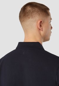 Marineblauwe polo met een gestructureerde stof, voorzien van een kraag en een klein, geborduurd logo op de achterkant. Minimalistisch ontwerp.