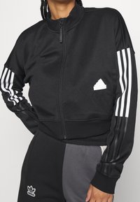 Svart zip-up träningsjacka med vit Adidas-logotyp och tre vita ränder på varje ärm. Har en hög krage och en mjuk tygstruktur.