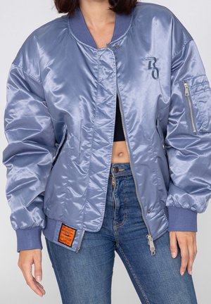 Vrouw draagt een glanzende lichtblauwe bomberjas over een zwart cropped top, gecombineerd met een blauwe spijkerbroek, staand tegen een effen achtergrond.