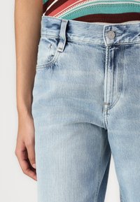 Light blue denim jeans feature a mid-rise waist, top button, and five-pocket design with subtle fading and visible stitching details.