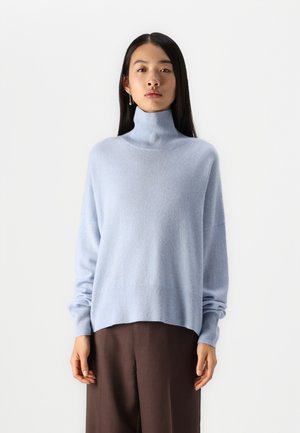 Hellblaues Rollkragensweatshirt aus weichem Strickmaterial, lockere Passform, überschnittene Schultern und gerippte Bündchen. Kombiniert mit braunen, weiten Hosen.