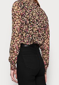 Blusa nera con motivo floreale, maniche lunghe e gonfie con polsini arricciati; presenta una combinazione di fiori rosa, gialli e bianchi su un tessuto trasparente.