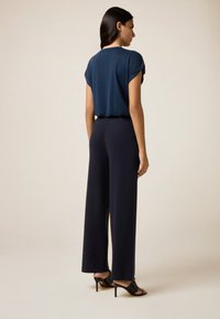 Vrouw die met de rug naar de camera staat, gekleed in een marineblauw T-shirt met korte mouwen, donkere wijde broek en zwarte hakken tegen een effen achtergrond.
