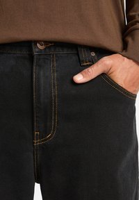 La mano di un uomo nella tasca anteriore di jeans neri con cuciture arancioni, indossando un maglione marrone.