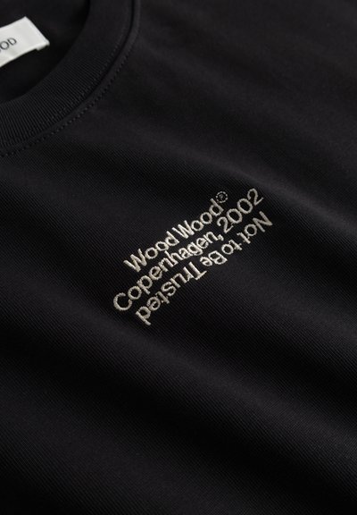 Μαύρο T-shirt με λευκό κεντημένο κείμενο "Wood Wood Copenhagen 2002 Not to be trusted" κοντά στο στήθος.