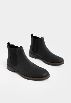 Bottines en cuir noir avec panneaux élastiques sur les côtés, bouts ronds et talon bas empilé. Présentent des accents de surpiqûres et des languettes pour un enfilage facile.