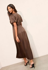 Rochie midi maro din satin, cu mâneci bufante, talie strânsă și fustă fluidă. Purtată cu sandale negre cu barete, evidențiind o textură fină.