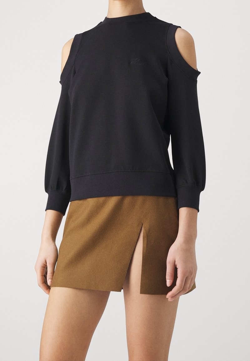 Sudadera negra de punto con cortes en los hombros, puños acanalados y una textura suave, combinada con una falda marrón con una abertura frontal.