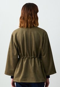 Cappotto verde oliva con cintura, realizzato in un tessuto morbido simile alla pelle scamosciata, maniche ampie e vestibilità comoda, che mostra dettagli di cucitura e una vita sfiancata.