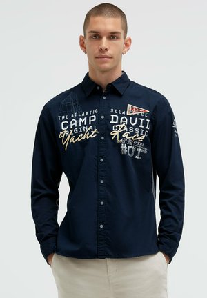 Junger Mann mit kurzen Haaren, trägt ein dunkelblaues Hemd mit Knöpfen mit weißem und goldenem Schriftzug zum Thema Yacht und hellbeige Hose.