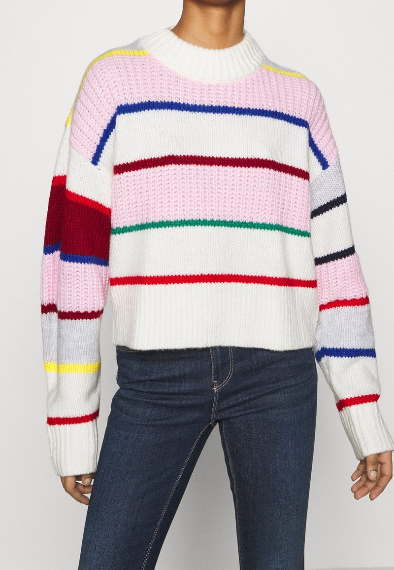Personne portant un pull en grosse maille avec des rayures horizontales multicolores associé à un jean bleu foncé.