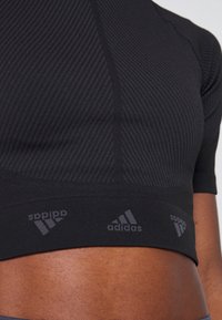 Gros plan d'une personne portant un crop top noir texturé Adidas avec des manches courtes et des bandes logo visibles au-dessus de la taille.