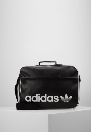 Musta suorakaiteen muotoinen Adidas olkalaukku, jossa valkoinen logo, vetoketjukiinnitys, ylhäällä kahva ja säädettävä olkahihna, lepää valkoisella pinnalla.