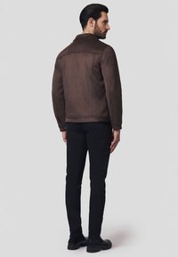 Brązowa zamszowa kurtka z zapięciem na napy, dwiema bocznymi kieszeniami i klasycznym kołnierzem, połączona z czarnymi dopasowanymi spodniami i butami.