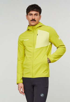 Mann mit Schnurrbart, der eine limettengrüne Reißverschlussjacke mit einer gelben Brusttasche und schwarzen Hosen vor einem schlichten grauen Hintergrund trägt.
