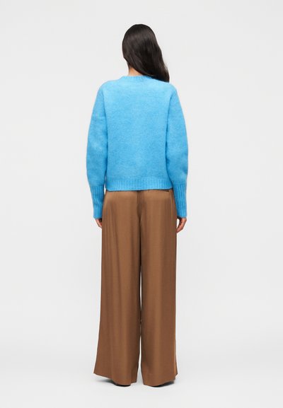 Γυναίκα με μακριά σκούρα μαλλιά που φοράει ένα φωτεινό μπλε πουλόβερ και φαρδιά καφέ παντελόνια, στέκεται με την πλάτη στραμμένη σε ένα απλό φόντο.
