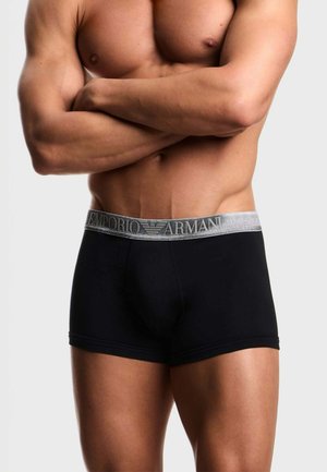 Torso maschile muscoloso con le braccia incrociate, che indossa boxer Emporio Armani neri con fascia in vita argentata.
