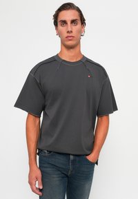 Young man with short wavy hair wearing a dark gray oversized T-shirt and blue jeans, standing with one hand in pocket against a white background.