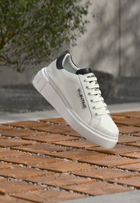 Baskets blanches Valentino avec talon noir et logo, présentées au-dessus d'un sol en bois et galets en plein air.