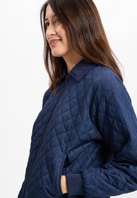 Marineblaues, gesteppte Bomberjacke mit einem gerippten Kragen, Reißverschluss und Seitentaschen, die das strukturierte Material und die taillierte Passform hervorhebt.