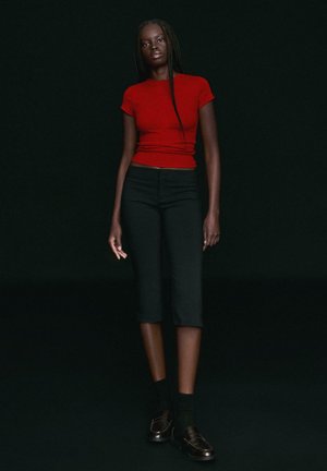 Frau mit langen Zöpfen, die ein eng anliegendes rotes Kurzarmtop, schwarze knöchellange Hose, schwarze Socken und schwarze Loafers trägt, steht vor einem dunklen Hintergrund.