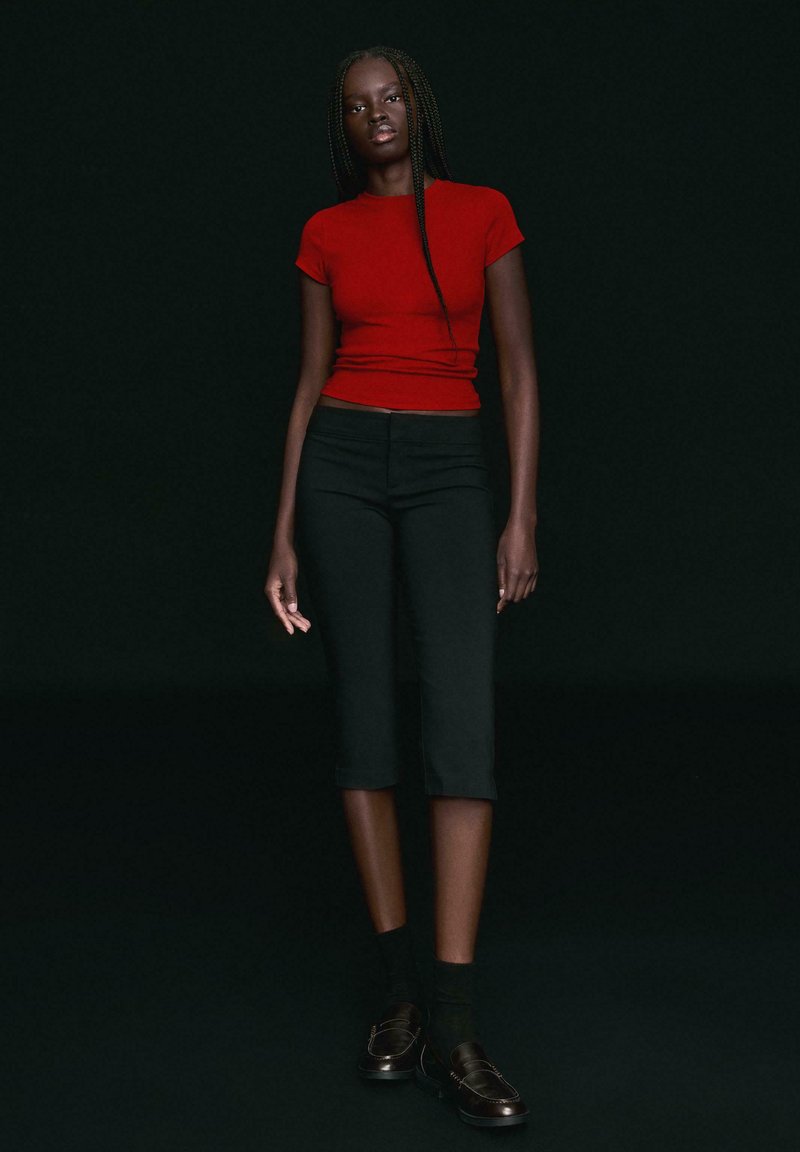 Žena s dlhými vrkočmi, oblečená v priliehavom červenom tričku s krátkym rukávom, čiernych krotovaných nohaviciach, čiernych ponožkách a čiernych mokasínach, stojí na tmavom pozadí.