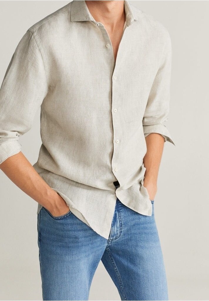 Homme portant une chemise en lin beige clair à manches longues retroussées et un jean bleu, mains partiellement dans les poches, sur un fond neutre.