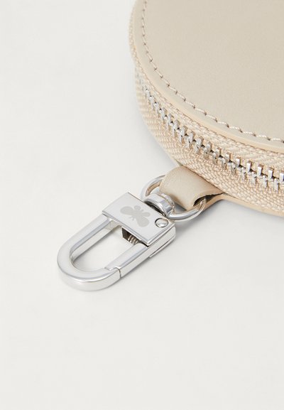 Estuche circular beige con una textura de cuero suave, que presenta un cierre metálico plateado y un gancho desmontable con un emblema de mariposa.
