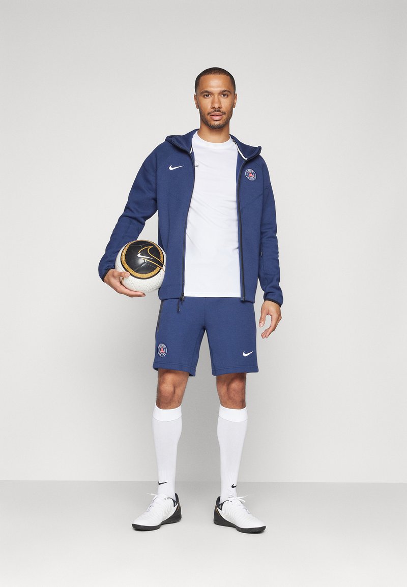 Completo sportivo Nike blu composto da una felpa con zip, t-shirt bianca, pantaloni corti e calze alte fino al ginocchio. Il modello tiene in mano un pallone da calcio bianco e nero.