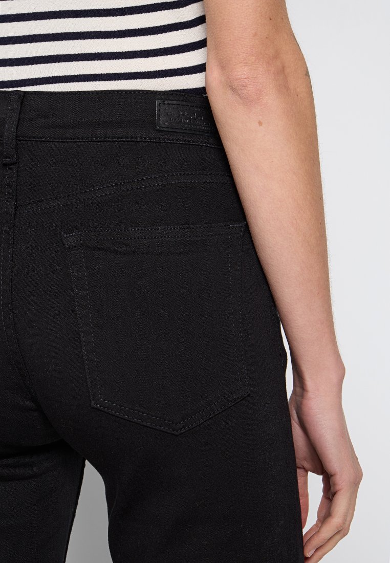 Rechter Hüfte und Arm einer Person, die schwarze Jeans und ein navy-blaues und weiß gestreiftes Hemd trägt, vor einem weißen Hintergrund.