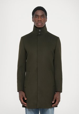 Manteau en laine vert foncé avec col haut, fermeture à boutons sur le devant et poches sur les côtés. La coupe est ajustée et légèrement allongée.