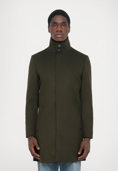 Manteau en laine vert foncé avec col haut, fermeture à boutons sur le devant et poches sur les côtés. La coupe est ajustée et légèrement allongée.