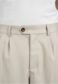 Pantalon beige taille haute avec un bouton marron foncé et des passants de ceinture, porté avec un haut blanc rentré.