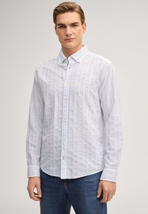 Jonge man die een lichtblauw overhemd met patroon en een blauwe spijkerbroek draagt, staand tegen een effen, neutrale achtergrond.