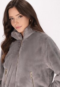 Faux fur grå jakke med en høj krave, guld lynlåsdetaljer og sidelommer. Blød tekstur og afslappet silhuet.