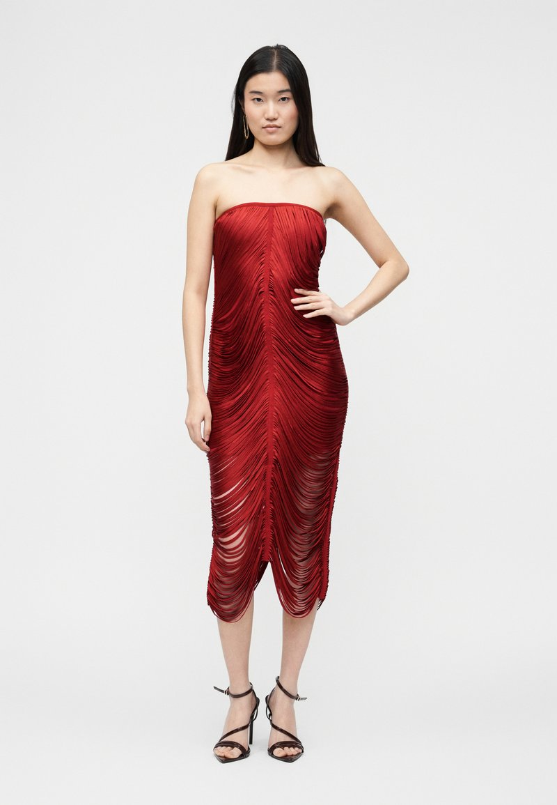 Femme portant une robe midi rouge froncée sans bretelles avec des sandales à talons hauts noires, debout devant un fond clair uni.